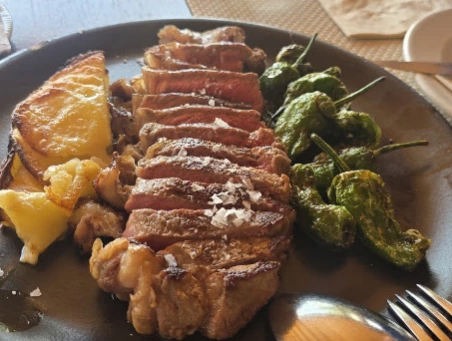 Corte de carne a la brasa laminado con sal en escamas, pimientos de padrón y papa, en plato de barro, en Casa Marcos